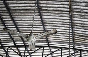 Old rusty gray metal ceiling fan hang on warehouse ceiling which covered with insulator to reduce temperature.